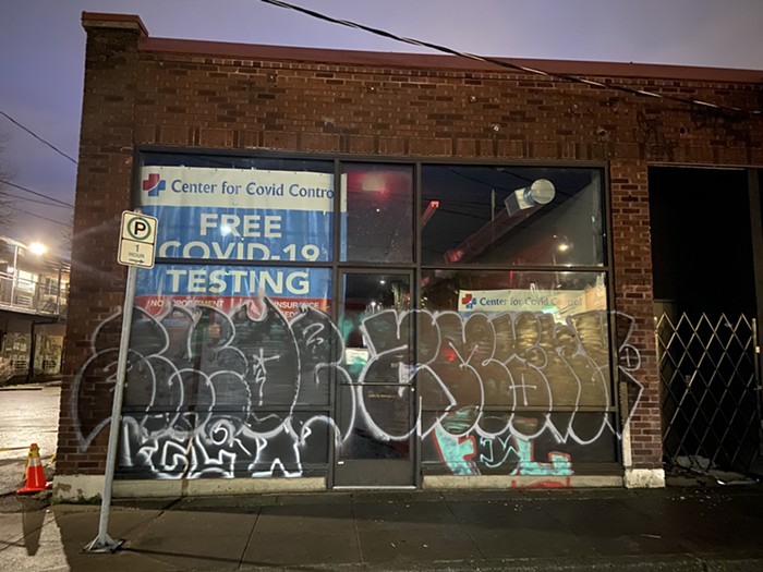 A brick building. The windows are tagged with spray paint. Behind the paint, you can see the tops of signs for covid testing